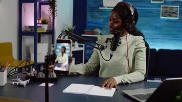 Smiling influencer african woman sitting in front of phone camera recording for blog. Digital blogger vlogger streaming talk show in studio using headphones, professional podcast microphone photo
