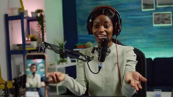 Focused shot on smartphone recording african blogger woman talking looking at camera during online podcast. Content creator streaming online broadcast, blogger discussing wearing headphones. photo