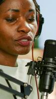 Close up of afro woman vlogger making new blog in home studio talking in professional microphone with audience. Speaking during livestreaming, blogger discussing in podcast wearing headphones. photo