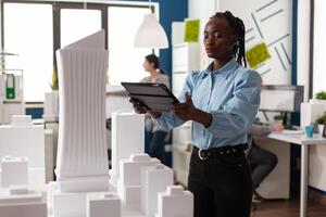 Architect woman of african american ethnicity working on tablet looking at professional maquette building model. Architectural worker viewing design for modern project in development photo