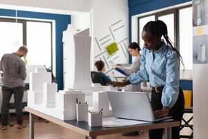 Architect african american woman working on laptop at workplace. Worker looking at building model maquette design for construction development. Modern urban layout project photo