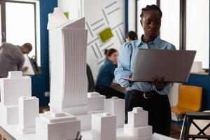 African american architect at business workplace looking at laptop and blueprint plan for building model maquette. Engineer constructor working on modern layout design for project photo
