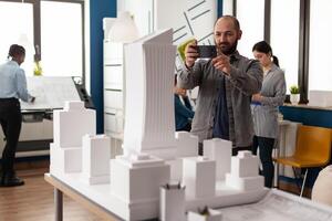 Architect profession man looking at maquette layout using smartphone to design building model plan maquette. Urban designer constructor working on professional modern construction photo