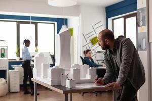 Man working on architectural design at office while looking at laptop computer for industry construction. Project manager inspecting maquette modern design building model photo