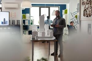 Architecture businessman working on tablet at office for designing building model maquette. Young builder inspecting development project sketch for modern structure technology photo