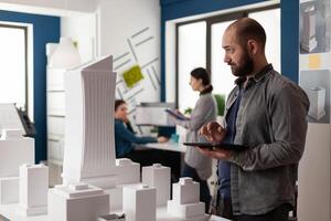 Man with architect profession working at office on building model design maquette on table. Caucasian architectural builder designing project of industrial construction photo