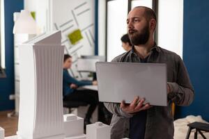 Professional man working as architect at office while using laptop computer for blueprint design. Adult constructor inspecting maquette plan bulding model for environment urban structure photo