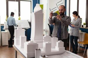 Architect profession man looking at maquette layout using smartphone to design building model plan maquette. Urban designer constructor working on professional modern construction photo
