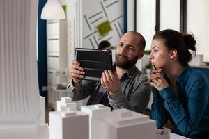 Design architecture colleagues sitting at office working on tablet and blueprints for layout plan maquette building model. Professional engineering team sitting for construction project photo