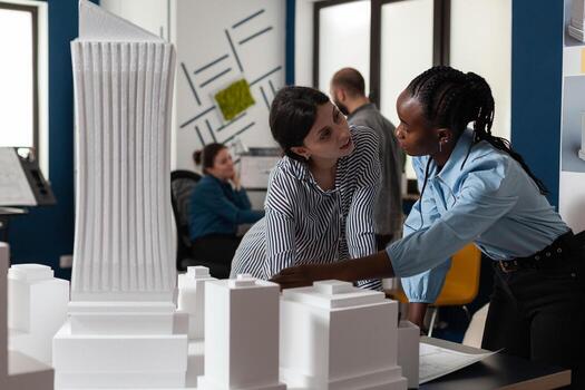Professional architecture multi ethnic women working on blueprints and building model maquette construction design. Team of workers pointing at architectural paper plan as occupation photo