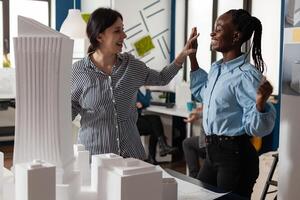 Architecture multi ethnic workmates in partnership to design building model maquette. Diverse professional engineer women highfive after success in blueprint plans cooperation photo