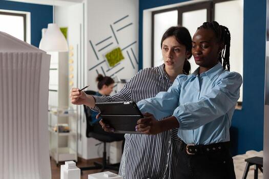 Diverse work team of multi ethnic architects working on tablet checking blueprint plan of building model maquette. Women analyzing project design on paper discussing construction photo