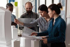 Team of diverse architects discussing blueprint plans on paper laptop computer tablet. Professional workers analyzing maquette building model for project design and construction photo