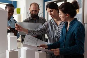 Team of diverse architects discussing blueprint plans on paper laptop computer tablet. Professional workers analyzing maquette building model for project design and construction photo