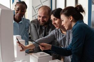 Multi ethnic architectural teamwork analyzing maquette building model construction layout. Diverse professional group of colleagues pointing at project design blueprint plan photo