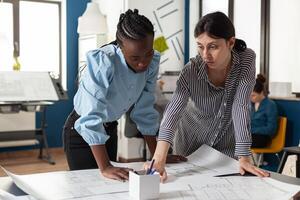 Architectural team of multi ethnic women plan sketch blueprint layout for design project building model maquette. Diverse group pointing at construction on paper for job occupation photo