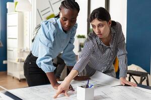 Architectural team of multi ethnic women plan sketch blueprint layout for design project building model maquette. Diverse group pointing at construction on paper for job occupation photo