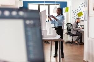 Architect working with smartphone standing next to table with white foam scale model in architectural modern office. Engineer with mobile device photographing maquette of real estate project. photo