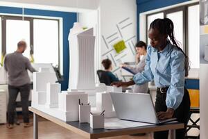 Standing architect with laptop looking at scale foam model of building from residential project. Architectural engineer comparing computer blueprints with 3d maquette of office complex development. photo
