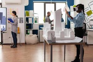 Architect standing in modern architectural office using virtual reality goggles to view 3d plan of white foam maquette. Engineer working with vr headest next to table with model of urban project. photo