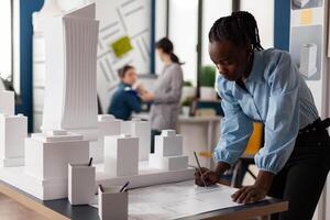 Residential architect drawing blueprints at desk in architectural office next to white foam scale model of skyscraper. Development engineer making notes on construction plan. photo