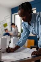 Portrait of architect making notes on construction plan papers at desk in modern architectural office. Development engineer drawing blueprints for residential project next to foam maquette. photo
