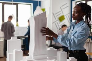 Architect standing at desk with white foam maquette of construction project in modern architectural office. Engineer inspecting futuristic 3d building design of skyscraper. photo