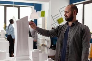 Inspired architect holding pen measuring skyscraper foam model standing next to desk with residential maquette. Engineer looking for inspiration in modern architectural office. photo