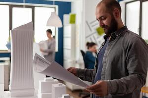 Architect reading blueprint of residential project next to skyscraper maquette in modern architectural office. Engineer inspecting construction plans standing near desk with 3d foam model. photo