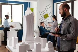 Architect holding tablet with blueprints inspecting design of white foam building model in architectural office. Engineer comparing construction plans with skyscraper maquette. photo