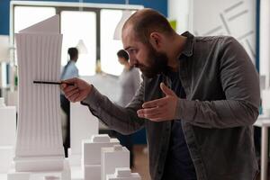 Engineer holding pen inspecting the design of skyscraper maquette in modern office. Inspired architect looking at the architectural lines of white foam model of residential project. photo