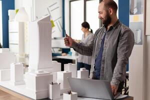 Inspired architect with laptop looking at the architectural lines of white foam model of residential project. Engineer holding pen inspecting the design of skyscraper maquette in modern office. photo