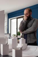 Architectural engineer thinking about design improvements standing next to desk with blueprints and architectural foam model. Architect looking for inspiration in modern office. photo