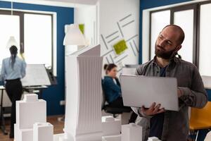 Architectural inspector holding laptop tilting head looking at maquette of skyscraper in residential project. Architect thinking about design improvements to urban planning scale model. photo