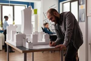 Architect drawing blueprints at desk in modern architectural office next to white foam scale model of skyscraper. Engineer making notes on construction plan leaning over desk. photo