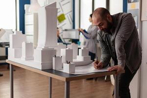 Development engineer making notes on construction plan of residential project leaning over desk with maquette. Architect drawing blueprints in architectural office next to white foam scale model. photo