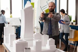 Architect working with smartphone to photograph white foam scale model maquette of skyscraper building complex. Engineer with mobile device next to desk in architectural modern office. photo