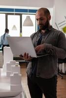 Architect holding laptop comparing blueprints with skyscraper maquette in architectural office. Engineer with portable computer looking at construction plans of real estate property. photo