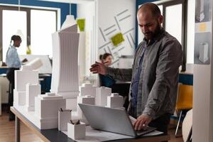 Arhitectural designer in videocall conference presenting white foam building model of residential buildings to client. Architect using laptop computer in modern office next to desk with 3d maquette. photo