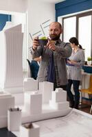 Smiling design architect holding smartphone to photograph desk with architectural model of skyscraper and buildings. Bearded engineer happy with final white foam maquette posting on social media. photo