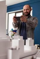 Professional architect holding smartphone smiling in videocall conference presenting final maquette to client. Bearded architectural engineer photographing table with white foam residential model. photo
