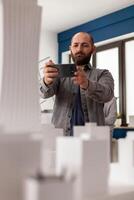Architect using smartphone to record of architectural maquette of skyscraper to post on social media. Bearded engineer photographing 3d white foam model with smart phone in modern office. photo