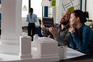Two architects working together looking at tablet with blueprints sitting at desk in modern architectural office. Project engineers doing teamwork using digital device next to table with scale model. photo