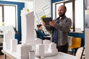 Bearded professional architect looking at tablet in front of white foam building model in architectural office. Engineer comparing blueprints on digital device with scale design of skyscraper. photo
