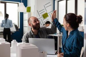 Two architects high fiving sitting at desk in architectural modern office with laptop and foam scale model of residential buildings. Successful happy smiling colleagues celebrating team project. photo