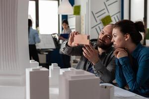 Architects doing teamwork looking at mobile content on smartphone sitting at desk in modern architectural office. Project engineers team collaboration using smart phone to photograph foam model. photo