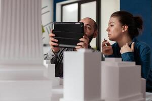 Project engineers working together looking at tablet with construction plans sitting at desk in modern architectural office. Two architects doing teamwork next to table with buildings maquette. photo