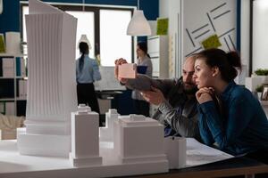 Team of two architect colleagues using smartphone in videocall conference with client presenting desk with buildings models. Architectural engineers working together with smart phone and maquette. photo