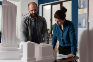 Architectural engineers doing teamwork reading construction plans standing next to table with foam model maquette. Team of two architects colleagues working together looking down at blueprins. photo