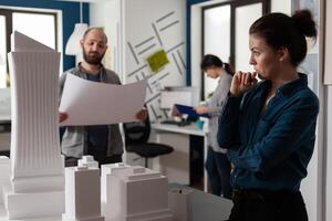 Architect looking into the distance gazing at skyscraper scale model with team colleague engineer holding construction plans. Team of architects brainstorming architectural ideeas. photo